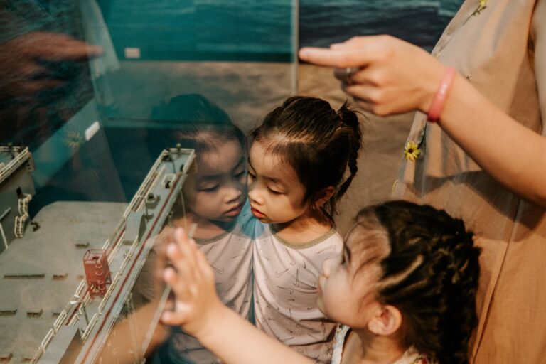 Two girls looking at a glass cabinet with the hand of an adult, clearly explaining what they can see. They look interested